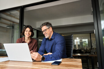 Male executive manager explaining corporate online project plan to female partner sitting at office desk. Two happy busy professionals business team people working together using laptop computer.