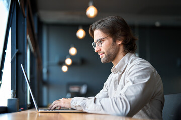 Fototapeta premium Busy young European business man entrepreneur looking at laptop at work. Businessman executive professional manager investor using computer sitting in office thinking of investment project plan.