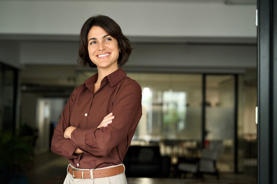 Happy confident young Hispanic professional business woman standing at work in office arms crossed looking away, proud businesswoman leader executive thinking of future career success.