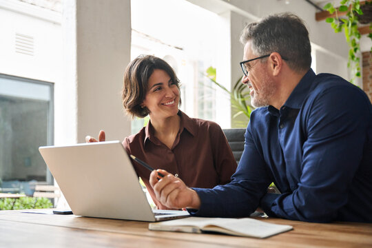 Two busy business team people using laptop computer working together sitting at office meeting. Happy young woman and mature man entrepreneurs or executives smiling at work talking in modern workspace