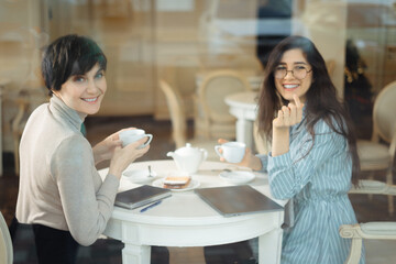 Two smiling girls have coffee time together in cafe, view through window glass