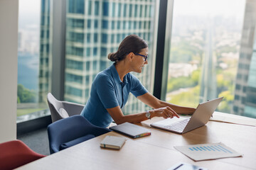 A determined and focused woman is using a laptop in a modern, sleek office that boasts impressive...