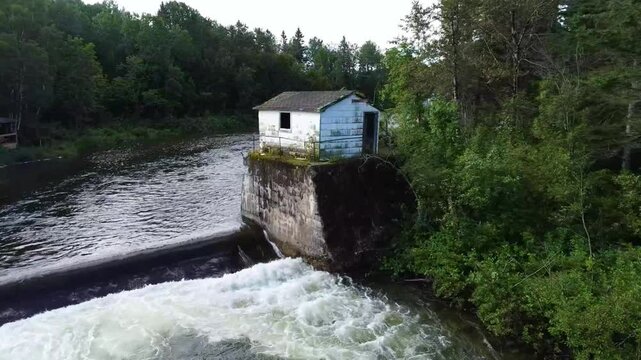 A drone flies up a old abandoned pump house cabin atop old cement foundations & its water intake along a salmon river surrounded by dense green forest. ( Mitis river, Qu&eacute;bec, Canada. )