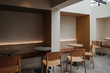 A serene, minimalist cafe interior featuring wooden chairs paired with round marble tables. The space has a warm and modern design with soft lighting, neutral tones, and a textured wall accent.