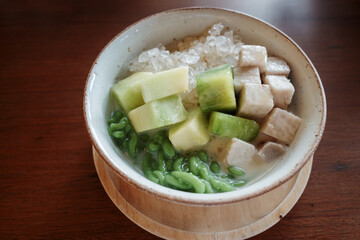 A close-up view of a refreshing traditional Thai dessert featuring a mix of diced taro, green pandan noodles, and melon pieces served in a bowl with coconut milk and crushed ice.