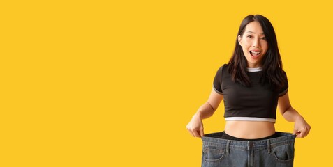 Happy young Asian woman in loose jeans on yellow background with space for text. Weight loss concept