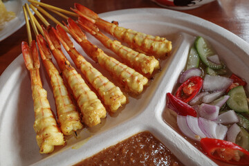 A close-up shot of delicious grilled satay shrimp skewers served with a side of peanut sauce and a fresh cucumber salad garnished with red chili peppers and sliced onions. 