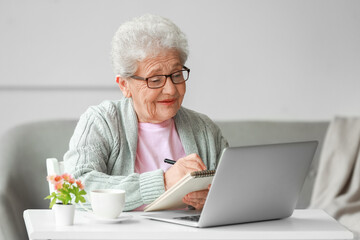 Senior woman with laptop writing in notebook at home