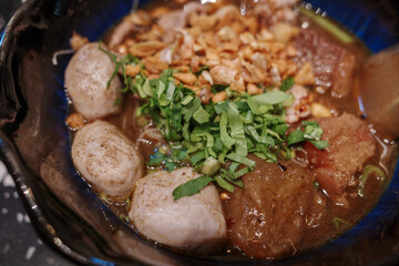 A close-up view of a traditional Thai boat noodle soup served in a black bowl, featuring tender pork meatballs, sliced pork, crispy fried garlic, and fresh herbs on top.