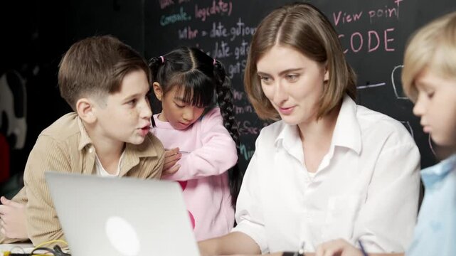 Caucasian teacher using laptop and explain about engineering system while diverse student holding electronic equipment in front of blackboard with prompt or code written in STEM class. Erudition.