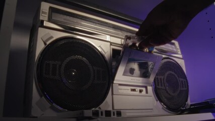A close-up shot of hand placing a cassette tape into a retro music player and pressing the play button. The neon lights in the studio create a vibrant, nostalgic atmosphere.