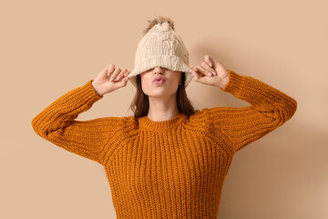 Young woman in winter clothes blowing kiss on beige background