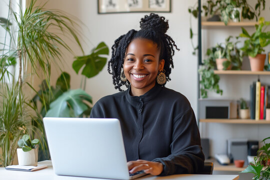 A smiling African American woman working remotely on a laptop with no logo at a desk in a well-lit, organized office space, with a few plants and shelves in the background