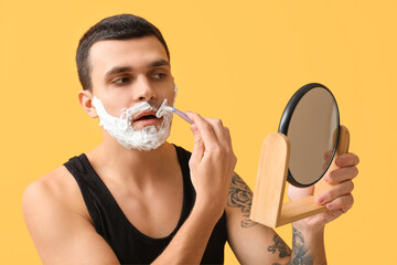Handsome young man with mirror and razor shaving his face on yellow background