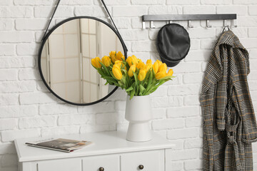 Vase of yellow tulips on dresser with mirror, hanger and coat near white brick wall in hallway
