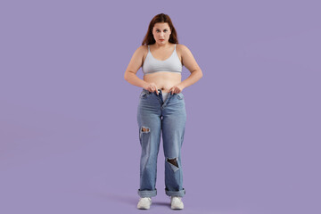 Overweight young woman in tight jeans on purple background. Weight gain concept