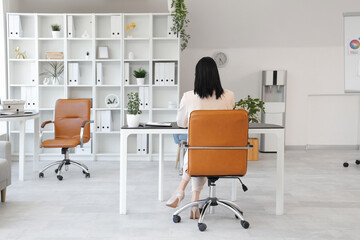 Young woman using modern laptop and sitting at workplace in light office