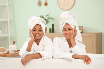 Happy young African-American mother and her daughter in bathrobes sitting inside bathtub at home