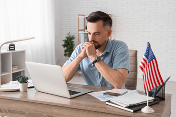Handsome young man with laptop and flag of USA working at table in office