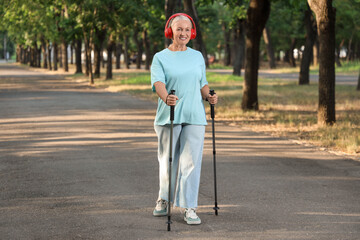 Mature woman in headphones training with walking poles in park