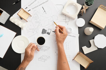 Male furniture designer with cup of coffee drawing on black table, top view