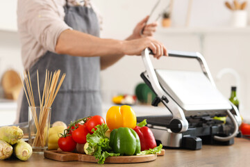 Young man cooking tasty fresh vegetables on modern electric grill in kitchen, closeup
