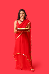 Fototapeta premium Beautiful young happy Indian woman in sari holding plate with candles and flowers on red background. Divaly celebration