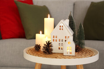 Burning candles with house shaped holders and Christmas decor on table in living room, closeup