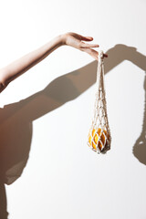 Person holding an orange in front of a white wall with a net bag hanging from their hand, casting a shadow