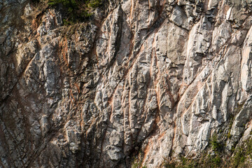 A close-up of a rocky cliff face, showcasing intricate textures and natural patterns formed over time. The sun illuminates the grooves and ridges, highlighting the rugged and weathered surface