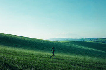 a woman jogging through lush green hills under a clear blue sky during the early morning in a serene landscape
