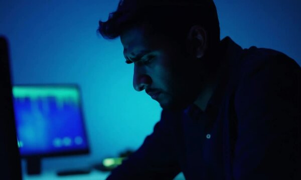 a man is frustrated in front his monitor in dark room. he is wiping his face and scratching his head.
