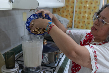 Mexican grandmother pours chopped walnuts into blender to prepare the nogada sauce for traditional...