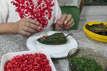 A woman in a red embroidered blouse stuffing a chile en nogada with ground beef in a home kitchen, preparing the iconic Mexican dish.