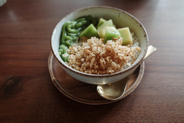 A close-up view of a refreshing traditional Thai dessert featuring a mix of diced taro, green pandan noodles, and melon pieces served in a bowl with coconut milk and crushed ice.