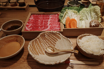 A beautifully arranged Shabu-Shabu meal featuring thinly sliced marbled beef, fresh vegetables including napa cabbage, carrots, and mushrooms, alongside bowls of dipping sauces and steamed rice.
