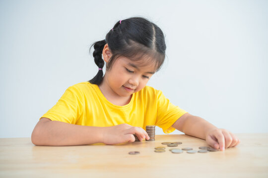 Asian young girl is sitting at a table with a pile of coins in front of her. She is focused on counting the coins and seems to be enjoying the activity