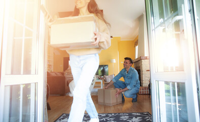 A couple is moving into a new home, carrying boxes through the open door. The sunlight pours in,...