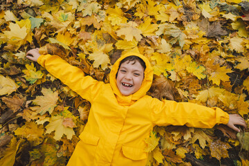 A child wearing a yellow raincoat lies happily among a in golden autumn leaves, smiling and enjoying the crisp fall weather. The leaves create a vibrant backdrop, showcasing the beauty of the season