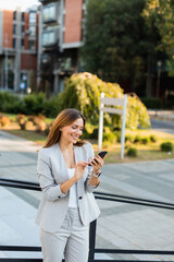 Young professional woman dressed in business attire smiles while texting on her smartphone in a vibrant urban setting during daylight