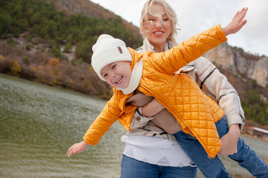 A beautiful young mother with kid spends time with her child outdoors together against the backdrop of a lake and mountains, the child frolic in the park. People dressed in warm autumn clothes