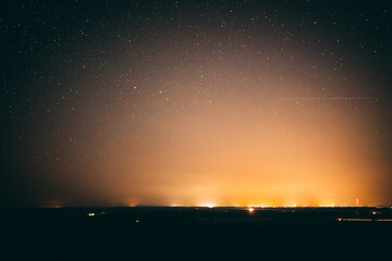 Fototapeta premium A serene night sky filled with stars stretches over a distant horizon illuminated by the soft glow of city lights. The contrast between the dark landscape and the warm light creates a peaceful