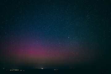 A breathtaking night sky filled with stars, highlighted by a faint aurora borealis in shades of pink and purple, illuminating the horizon above a quiet town. The scene captures the serene beauty 