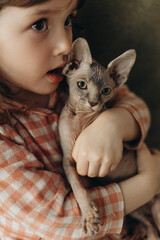 little cute girl 3 years old wearing pajamas hugging her canadian sphynx kitten sitting on a chair at home
