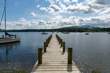 Obraz premium View over length of small boat pier into Lake Windemere in Lake District National Park near Ambleside, UK with sailboats in water against a white clouded blue sky