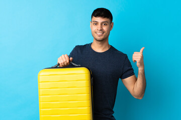 Young Colombian man isolated on blue background in vacation with travel suitcase and with thumb up