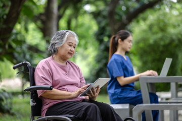 Obraz premium Elderly woman in a wheelchair using a tablet while a nurse works on a laptop nearby, enjoying a quiet moment in the park