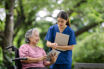 Elderly woman smiling as she uses a tablet, with a nurse standing by, observing and ready to assist in a peaceful park setting.