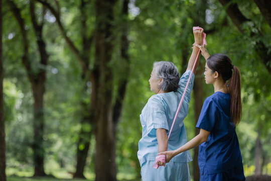 Nurse assisting an elderly woman with a resistance band exercise in a peaceful outdoor park, promoting strength and wellness.