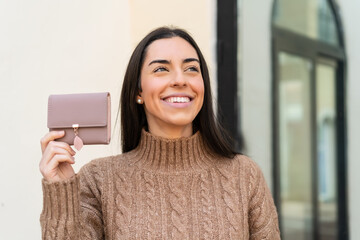 Young woman holding a wallet at outdoors looking up while smiling